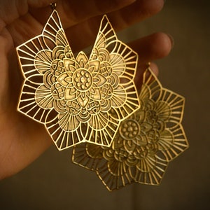 K&ouml;nnte beinhalten: Ein Paar goldfarbene Ohrringe aus Metall mit einem filigranen, komplizierten Mandala-Design. Die Ohrringe sind gro&szlig; und haben eine runde Form mit einem floralen Muster.