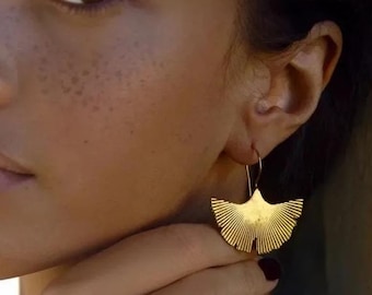 May include: A woman with dark brown hair and freckles wears gold earrings shaped like a fan. The earrings are dangling and have a textured surface.