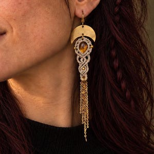 May include: A long dangle earring featuring a brass crescent moon shape at the top, a central oval amber-colored stone encased in light-colored macrame cord, and a series of delicate gold-toned metal chains hanging from the bottom of the piece.