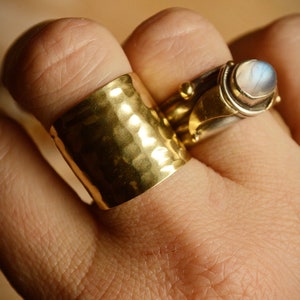 May include: Two gold rings on a finger. The ring on the index finger is a wide band with a hammered texture. The ring on the middle finger is a silver band with a moonstone set in a gold bezel.