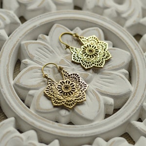 K&ouml;nnte beinhalten: Ein Paar goldfarbene Filigran-Ohrringe mit floralem Design. Die Ohrringe werden auf einer wei&szlig;en, geschnitzten Holzoberfl&auml;che mit floralem Muster pr&auml;sentiert. Die Ohrringe haben einen Hakenverschluss.