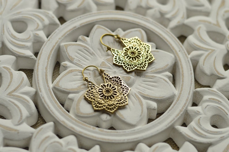 K&ouml;nnte beinhalten: Ein Paar goldfarbene Filigran-Ohrringe mit floralem Design. Die Ohrringe werden auf einer wei&szlig;en, geschnitzten Holzoberfl&auml;che mit floralem Muster pr&auml;sentiert. Die Ohrringe haben einen Hakenverschluss.