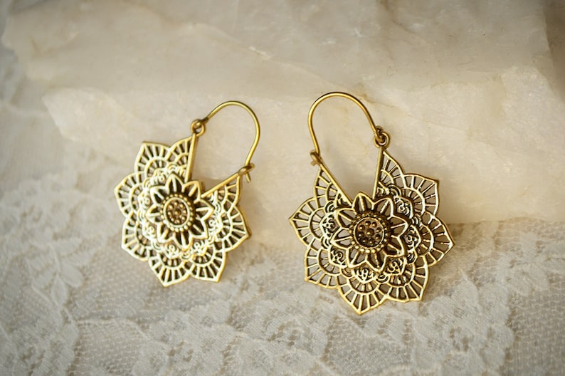 K&ouml;nnte beinhalten: Ein Paar goldfarbene, durchbrochene Ohrringe. Jeder Ohrring hat ein florales Design mit komplizierten Details. Die Ohrringe haben einen Hakenverschluss und werden auf einer wei&szlig;en, strukturierten Oberfl&auml;che pr&auml;sentiert.