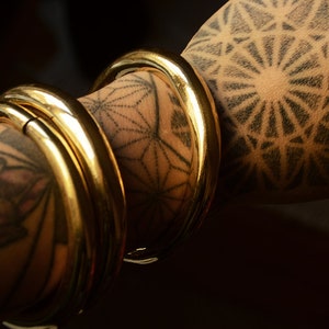 May include: Close-up of several thick, gold bangles stacked on an arm. The arm is decorated with intricate black tattoos, including geometric patterns and floral designs. The bangles have a polished, reflective surface.