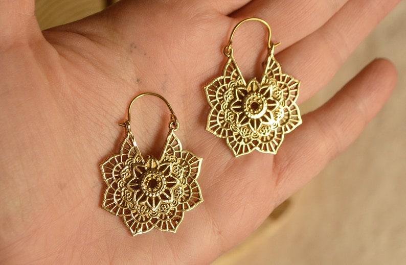 K&ouml;nnte beinhalten: Ein Paar goldfarbene Ohrringe mit einem floralen Mandala-Design. Die Ohrringe haben einen Klappverschluss und bestehen aus einem gl&auml;nzenden, metallischen Material. Das filigrane Openwork-Design zeigt eine zentrale Blume und umliegende Bl&uuml;tenbl&auml;tter.