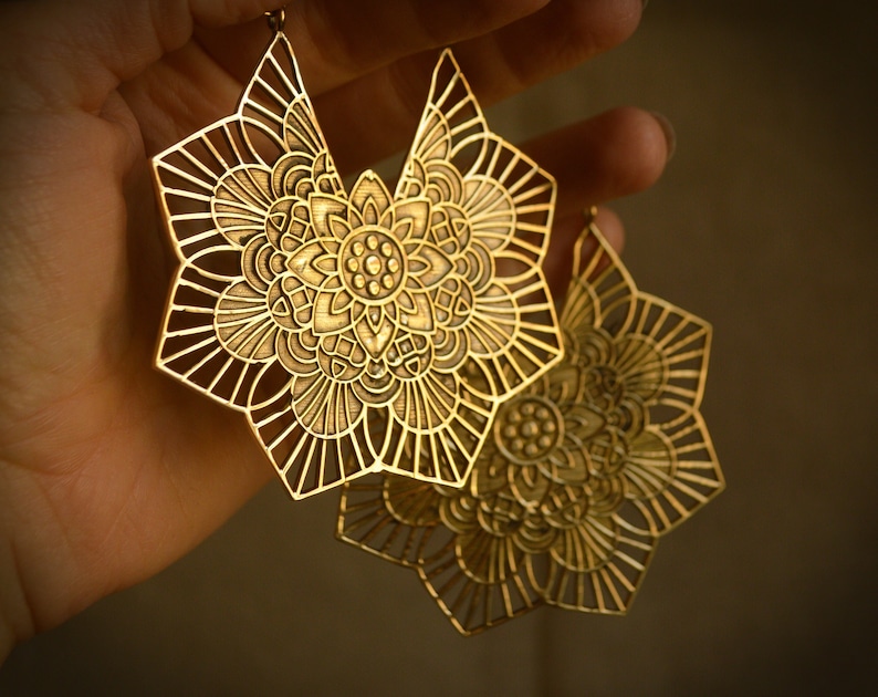 K&ouml;nnte beinhalten: Ein Paar goldfarbene Ohrringe aus Metall mit einem filigranen, komplizierten Mandala-Design. Die Ohrringe sind gro&szlig; und haben eine runde Form mit einem floralen Muster.