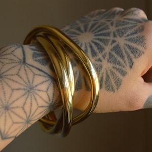 May include: Three gold-toned metal bangles stacked on a wrist. The bangles are smooth and shiny, and they are worn on a wrist with a geometric tattoo.