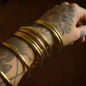 以下が含まれることがあります： タトゥーのある腕に、複数のゴールドのバングルが飾られています。バングルは重ねてつけられており、大胆で目を引くルックになっています。