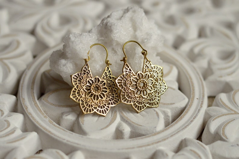 K&ouml;nnte beinhalten: Ein Paar goldfarbene, durchbrochene Ohrringe. Jeder Ohrring hat ein florales Design mit komplizierten Details. Die Ohrringe werden auf einer wei&szlig;en, verzierten Oberfl&auml;che mit einem wei&szlig;en Felsen dahinter pr&auml;sentiert.