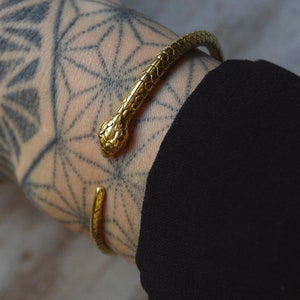 May include: A gold snake-shaped bangle bracelet with a textured surface. The bracelet is worn on a person's wrist.