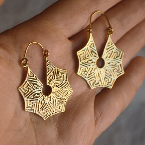 May include: A pair of gold-toned hoop earrings with a geometric design. The earrings are made of metal and have a circular shape with a cutout in the center.