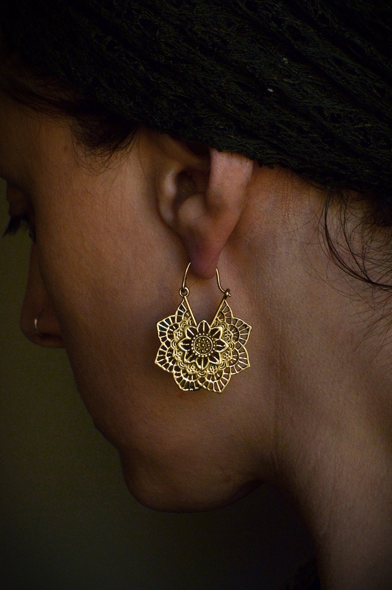 K&ouml;nnte beinhalten: Goldfarbene, durchbrochene Ohrringe mit floralem Design. Die Ohrringe haben die Form einer stilisierten Blume mit einem zentralen kreisf&ouml;rmigen Element und komplizierten Bl&uuml;tenblattdetails. Die Ohrringe h&auml;ngen an einem Haken.