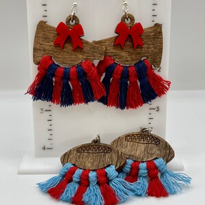 May include: Earrings with a football theme. Two pairs of earrings are displayed. One pair features a wooden megaphone shape with a red bow and red and blue tassels. The other pair has a wooden football shape with red and light blue tassels.