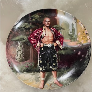 Könnte beinhalten: Dekorative Platte mit einem Gemälde eines Mannes in einem roten, floralen Gewand und schwarzen Shorts mit weißen floralen Akzenten. Der Hintergrund zeigt eine Gartenszene mit Statuen und Laub. Die Signatur des Künstlers, "William T. Chambers", ist sichtbar.