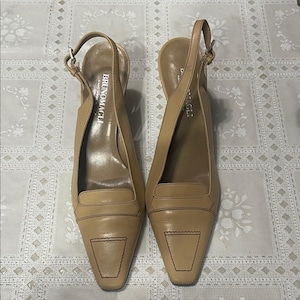 Puede incluir: Un par de zapatos de tacón slingback de cuero color beige con puntera. Los zapatos tienen un tacón pequeño, una correa en el tobillo con hebilla y costuras decorativas. La marca "BRUNOMAGLI" es visible en la plantilla.