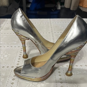 May include: A pair of silver peep-toe high heels. The heels have a unique design with a spiral of gold, copper, and silver-colored metal. The platform and heel base are also accented with gold and copper.