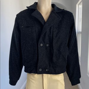 May include: A black double-breasted jacket with a textured appearance. The jacket features a notched collar, two front pockets with zippers, and a ribbed hem. The buttons are black, and the jacket is displayed on a mannequin.