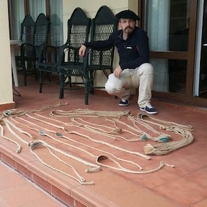 Könnte beinhalten: Ein Mann in einer schwarzen Jacke und einer Baskenmütze steht auf einer Veranda mit einem Satz geflochtener Seilschleudern. Die Schleudern liegen auf dem Holzboden der Veranda.