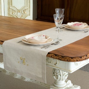 Puede incluir: Un camino de mesa de lino beige con un diseño bordado de un lazo y estrellas. El camino de mesa está colocado sobre una mesa de madera con una base pintada de blanco. Dos platos, un tazón y dos copas de vino están colocados sobre la mesa.