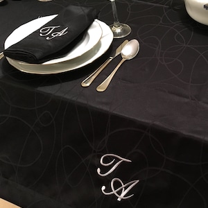Op de afbeelding: Een zwarte tafelkleed met een wit swirl-patroon. Twee zilveren vorken en een lepel liggen op een wit bord. Een zwarte servet met de initialen "T.A." geborduurd in wit is gevouwen op het bord. Het tafelkleed heeft de initialen "T.A." geborduurd in wit in de rechterbenedenhoek.