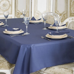 May include: A navy blue tablecloth is set on a rectangular table with white chairs. The table is set with white plates, bowls, and glasses. There are napkins folded into triangles on the plates.