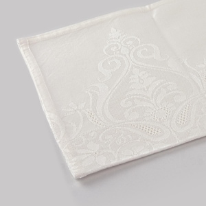 May include: Ivory-colored, folded linen napkin with an ornate, raised floral and scrollwork pattern along one edge. The fabric has a subtle sheen, and the design is detailed and elegant. The napkin's corner is visible, showing a clean, finished hem.