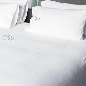 Può includere: Set di biancheria da letto bianco con iniziali ricamate. La biancheria da letto comprende due cuscini e un lenzuolo con angoli. Le iniziali sono di colore grigio argento. Il letto è ben fatto, con i cuscini disposti a capotesta.