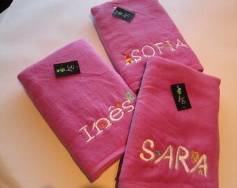 Op de afbeelding: Drie roze badhanddoeken met de geborduurde namen Sofia, Inês en Sara. Elke handdoek heeft een klein label. De namen zijn in witte draad geborduurd met bloemmotieven. De handdoeken zijn opgevouwen.