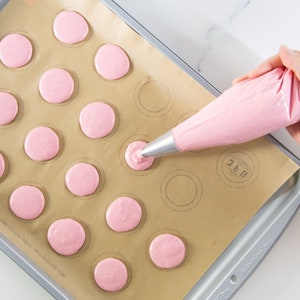 May include: A pink piping bag is being squeezed to fill circles on a baking sheet lined with parchment paper. The circles are outlined in black and the baking sheet has a silver rim.