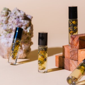 May include: Four glass bottles with black roll-on tops filled with a clear liquid and dried flowers. The bottles are arranged on a light beige surface with a stack of wooden blocks and a large white rock.