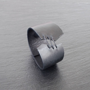 Op de afbeelding: Een zwarte leren manchetarmband met een uniek ontwerp. De armband heeft een brede band met een overlappend gedeelte dat met drie zichtbare steken aan elkaar is genaaid. De armband wordt getoond op een donkergrijze ondergrond.