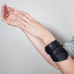 May include: A black leather bracelet with a unique, layered design. The bracelet is wrapped around a person's wrist.