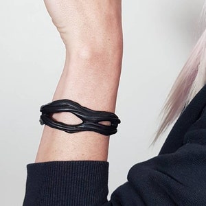 Könnte beinhalten: Ein mattes schwarzes Armband mit einem organischen, unregelmäßigen und wellenförmigen Design mit einer zentralen Öffnung. Das Stück hat ein strukturiertes, fließendes Aussehen, das sich um das Handgelenk legt und eine durchgehende Form zeigt.