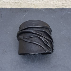 Puede incluir: Una pulsera brazalete ancha de cuero negro que presenta una serie de pliegues horizontales y texturas fruncidas en la superficie. El accesorio tiene un acabado mate liso y un dise&ntilde;o estructurado y abierto para la mu&ntilde;eca.