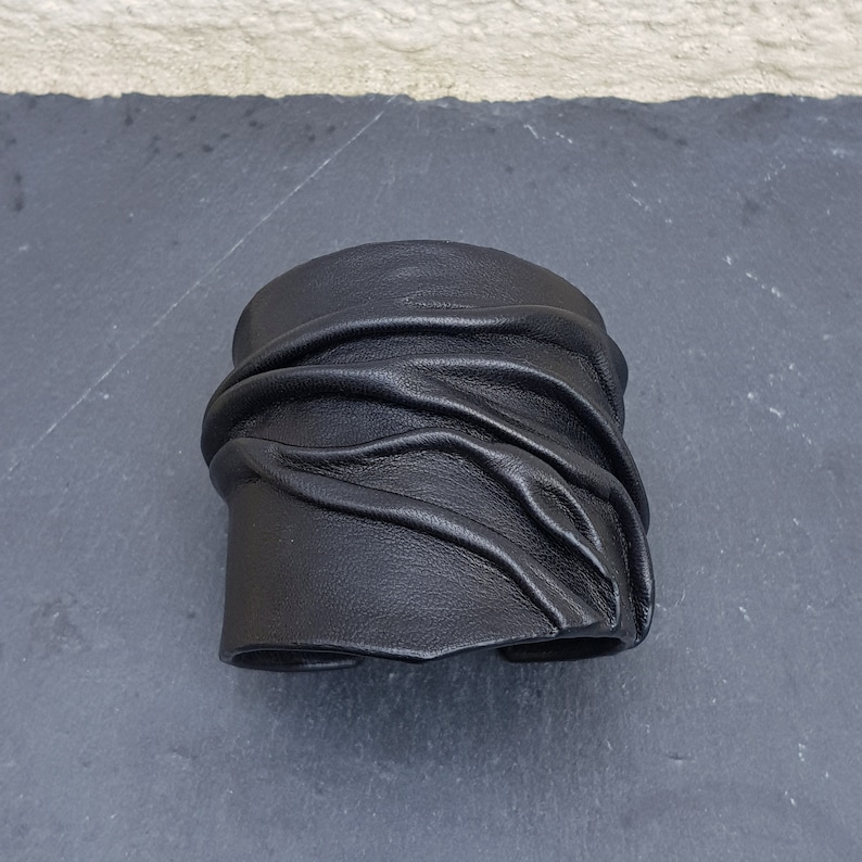 Puede incluir: Una pulsera brazalete ancha de cuero negro que presenta una serie de pliegues horizontales y texturas fruncidas en la superficie. El accesorio tiene un acabado mate liso y un dise&ntilde;o estructurado y abierto para la mu&ntilde;eca.