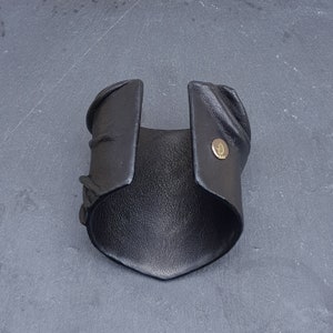 Puede incluir: Una pulsera brazalete de cuero negro ancha con superficie texturizada y borde puntiagudo. El brazalete cuenta con un broche de presi&oacute;n met&aacute;lico en tono dorado en un lado, que muestra un peque&ntilde;o logotipo circular grabado.