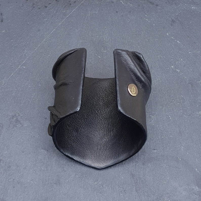 Puede incluir: Una pulsera brazalete de cuero negro ancha con superficie texturizada y borde puntiagudo. El brazalete cuenta con un broche de presi&oacute;n met&aacute;lico en tono dorado en un lado, que muestra un peque&ntilde;o logotipo circular grabado.