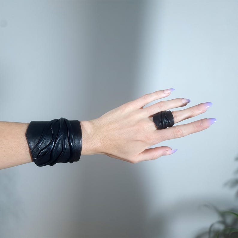 Puede incluir: Una pulsera de brazalete de cuero negro con una textura fruncida y un anillo de cuero negro a juego con un dise&ntilde;o fruncido similar. Ambas piezas est&aacute;n hechas de material oscuro y liso con pliegues y arrugas visibles.