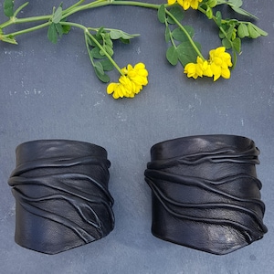 Puede incluir: Dos brazaletes de cuero negro con una serie de crestas horizontales, elevadas y onduladas en la superficie. Los brazaletes est&aacute;n colocados debajo de un tallo verde con peque&ntilde;as flores de color amarillo brillante.