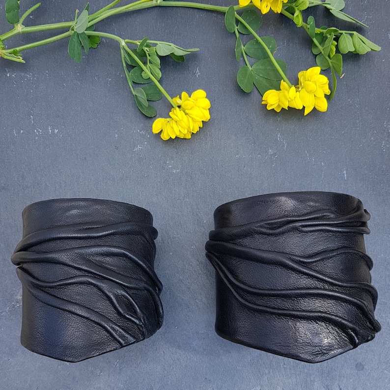 Puede incluir: Dos brazaletes de cuero negro con una serie de crestas horizontales, elevadas y onduladas en la superficie. Los brazaletes est&aacute;n colocados debajo de un tallo verde con peque&ntilde;as flores de color amarillo brillante.