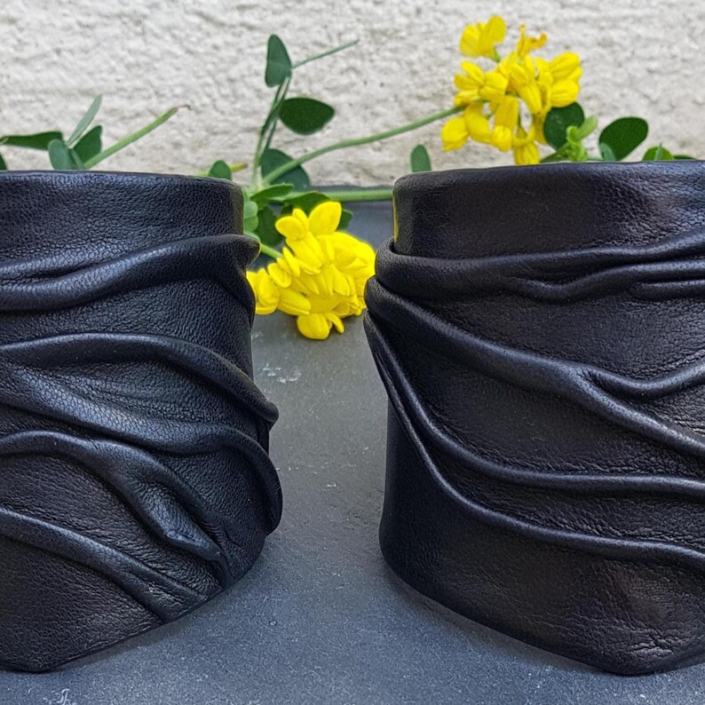 Puede incluir: Dos brazaletes de cuero negro con una superficie texturizada y fruncida con l&iacute;neas horizontales elevadas y fluidas. Los brazaletes tienen una forma cil&iacute;ndrica ancha y un acabado mate, presentados con flores amarillas de fondo.
