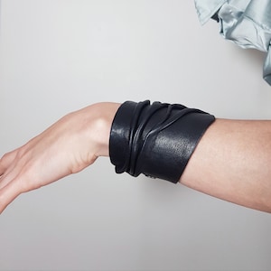 Puede incluir: Una pulsera brazalete ancha de cuero negro con una superficie texturizada que presenta m&uacute;ltiples pliegues y frunces suaves. La pulsera envuelve la mu&ntilde;eca y se asegura con un peque&ntilde;o cierre de presi&oacute;n met&aacute;lico visible en el lateral.