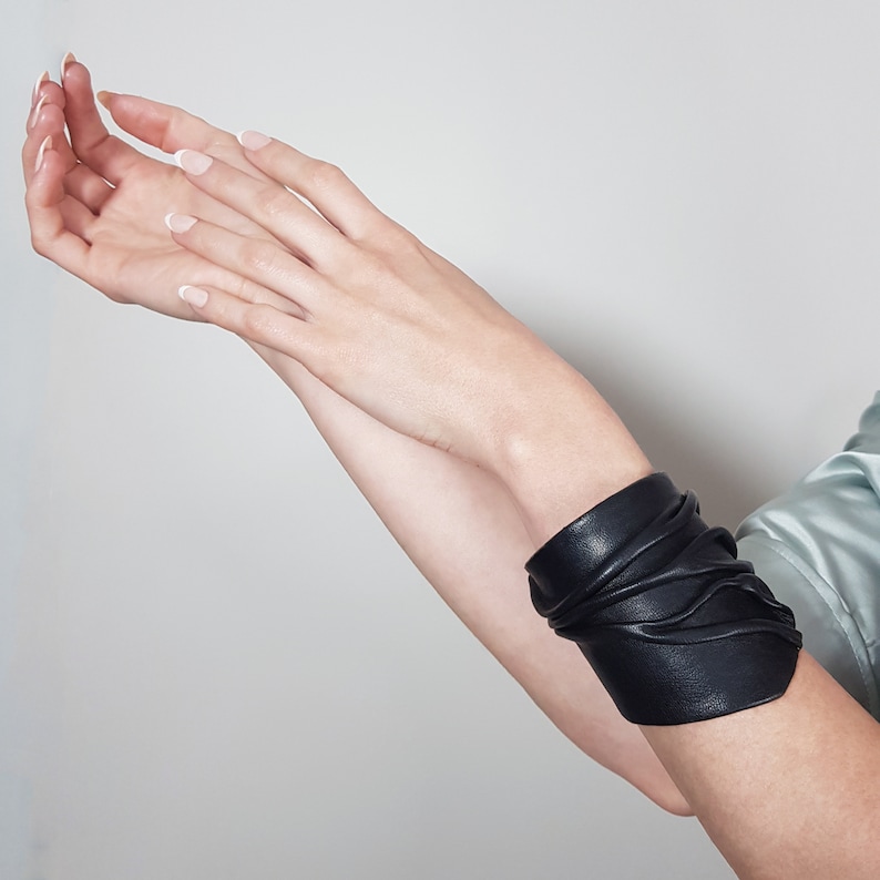 May include: A black leather bracelet with a unique, layered design. The bracelet is wrapped around a person's wrist.