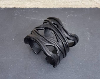 Puede incluir: Una pulsera tipo brazalete de color negro que presenta una serie de tiras superpuestas y sinuosas que crean un patrón abierto y orgánico. La superficie del material tiene un acabado mate con textura visible, formando una banda ancha y rígida.