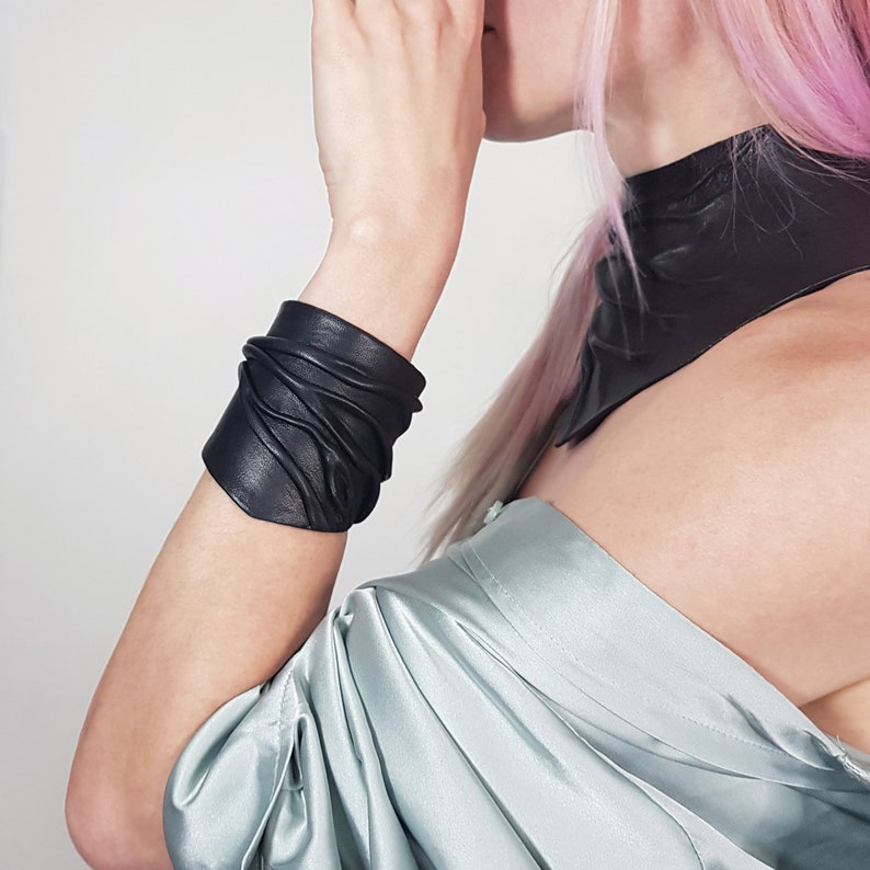 Puede incluir: Un brazalete de cuero negro con una textura fruncida y plisada. El accesorio se lleva en el antebrazo, combinado con un collar de cuero negro y una prenda de sat&eacute;n azul claro drapeada sobre la zona del hombro.