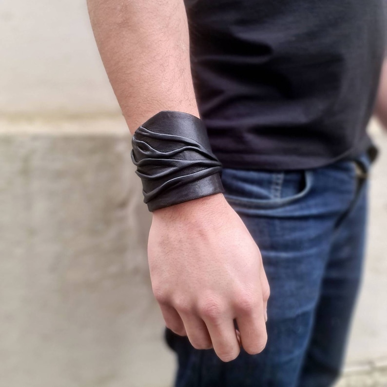 Puede incluir: Una pulsera ancha de cuero negro con una serie de pliegues horizontales fruncidos que crean una apariencia texturizada y superpuesta en la superficie de la banda, envolvi&eacute;ndose firmemente alrededor del antebrazo.
