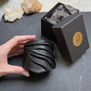 Puede incluir: Una pulsera brazalete de cuero negro ancha que presenta una serie de pliegues horizontales fruncidos en su superficie. La pulsera se presenta junto a una caja de regalo rectangular negra con una pegatina dorada circular con el texto O.b.s.o.