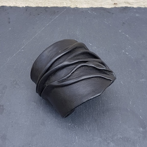 Puede incluir: Una pulsera brazalete ancha de cuero negro que presenta una serie de pliegues horizontales fruncidos que crean una superficie texturizada y ondulada en la parte frontal. El cuero tiene un acabado mate y grano visible.
