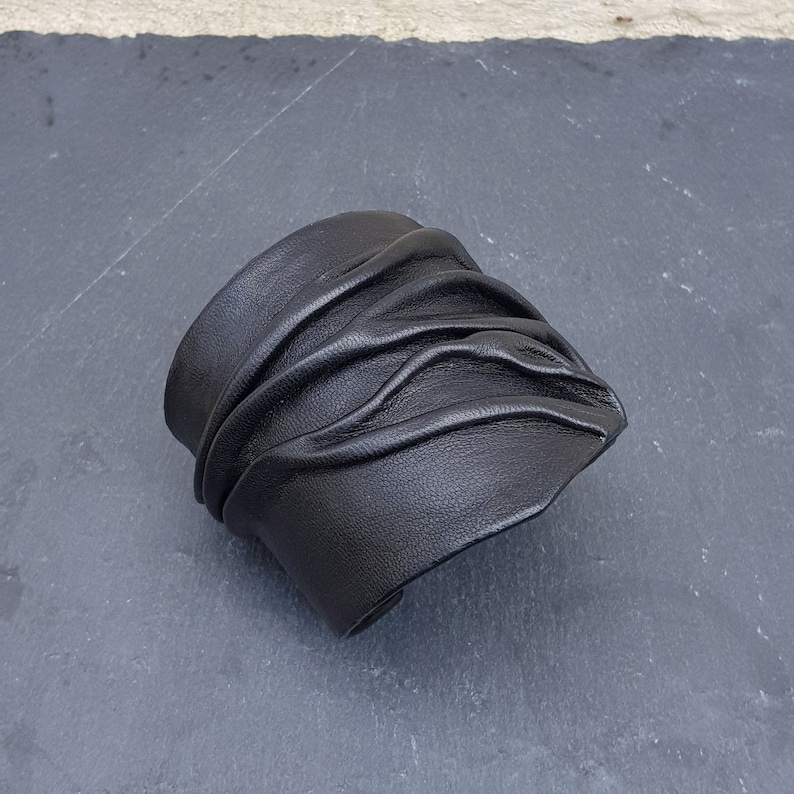 Puede incluir: Una pulsera brazalete ancha de cuero negro que presenta una serie de pliegues horizontales fruncidos que crean una superficie texturizada y ondulada en la parte frontal. El cuero tiene un acabado mate y grano visible.
