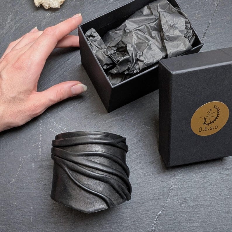 Puede incluir: Una pulsera de cuero negro con textura fruncida y superpuesta. La pulsera se muestra junto a una caja de regalo negra abierta que contiene papel de seda negro y una caja negra cerrada con una pegatina circular dorada con el texto O.b.s.o.